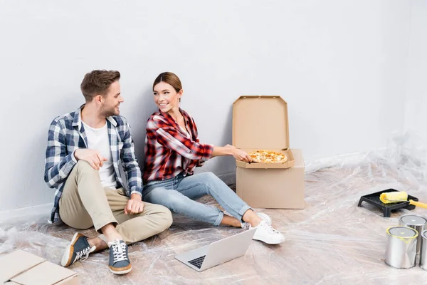 Piena lunghezza del sorriso giovane donna dando pezzo di pizza per l'uomo vicino computer portatile sul pavimento — Foto stock