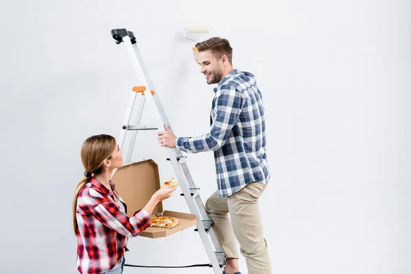 Sonriente joven con la pared de la pintura del rodillo mirando a mujer que da pedazo de pizza cerca de escalera aislada en gris - foto de stock
