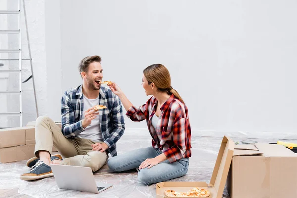 Полная длина счастливой женщины, кормящей мужчину куском пиццы, сидя на полу рядом с ноутбуком дома — стоковое фото