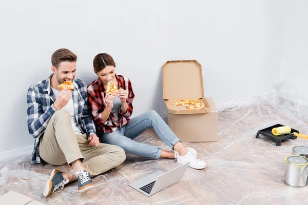 Vollständige Länge des jungen Paares Pizza essen, während es neben Laptop auf dem Boden zu Hause sitzt — Stockfoto