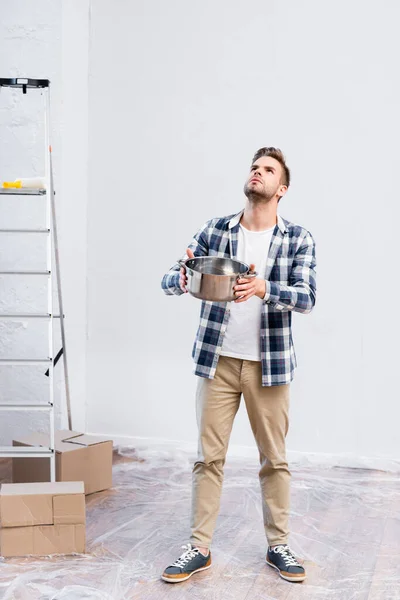 Voller Länge junger Mann mit Kochtopf, der in der Nähe von Pappkartons und Leiter unter undichter Decke zu Hause steht — Stockfoto