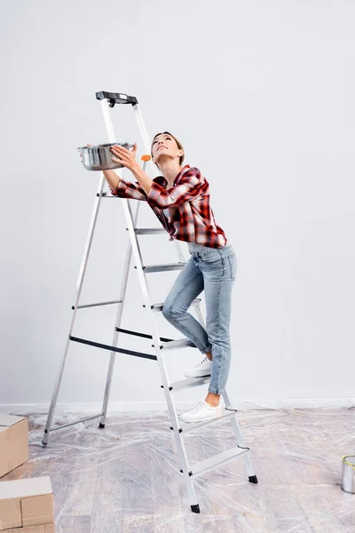 Volle Länge der jungen Frau, die nach oben schaut, während sie Topf auf Leiter unter undichter Decke zu Hause hält — Stockfoto