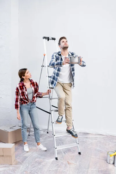 Pleine longueur de jeune homme tenant pot tout en se tenant sur l'échelle près de la femme sous le plafond fuyant à la maison — Photo de stock