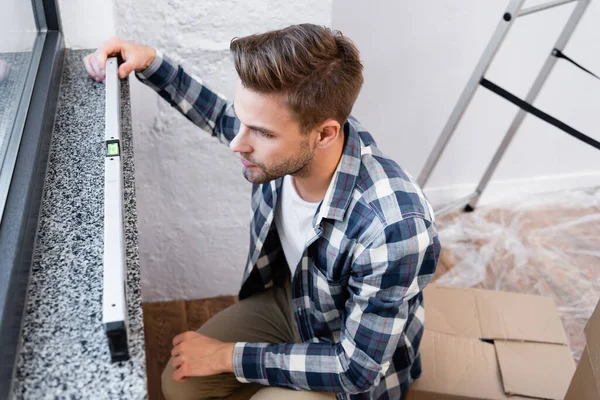 Vue grand angle du jeune homme tenant l'outil de mesure de niveau sur le rebord de la fenêtre sur fond flou à la maison — Photo de stock