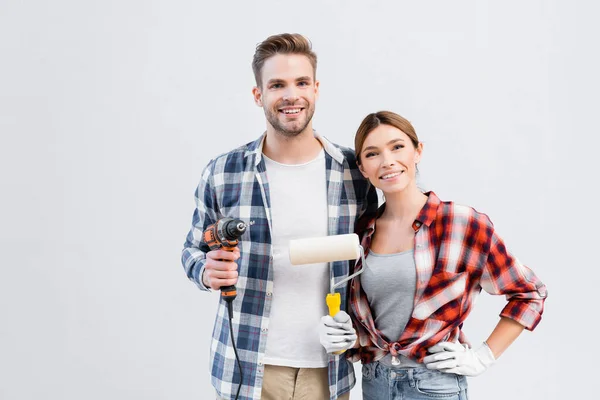 Glückliches junges Paar mit Bohrmaschine und Farbroller schaut in die Kamera, während es sich zu Hause umarmt — Stockfoto