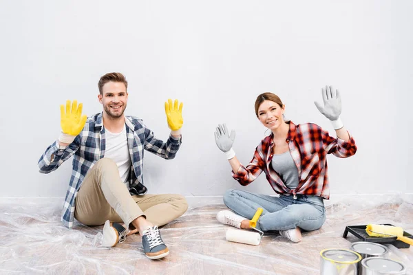 Full length of happy young couple looking at camera and showing gloves while sitting on floor at home — Stock Photo