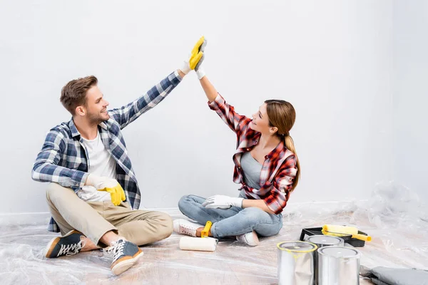 Longitud completa de feliz joven pareja en guantes dando unos a otros cinco altos mientras están sentados en el suelo cerca de latas de pintura en casa — Stock Photo