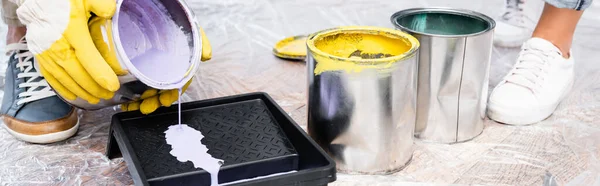Cropped view of man in gloves pouring purple paint from tin in roller tray on floor indoors, banner — Stock Photo