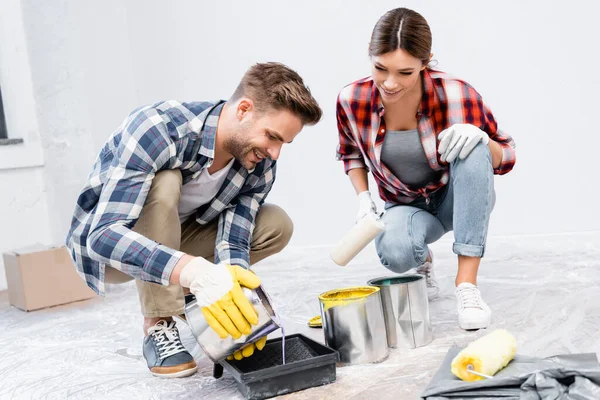 Longitud completa de sonriente joven vertiendo pintura en bandeja de rodillos cerca de la mujer en casa - foto de stock