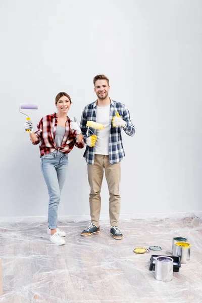 Longitud completa de feliz pareja joven con rodillos de pintura mirando a la cámara mientras muestra los pulgares hacia arriba en casa - foto de stock