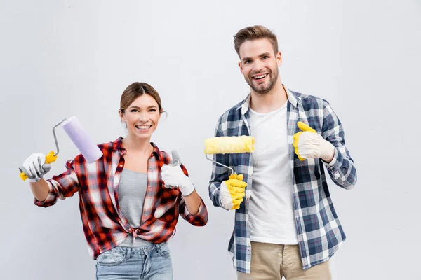 Feliz pareja joven con rodillos de pintura mostrando los pulgares hacia arriba mientras mira la cámara aislada en gris - foto de stock