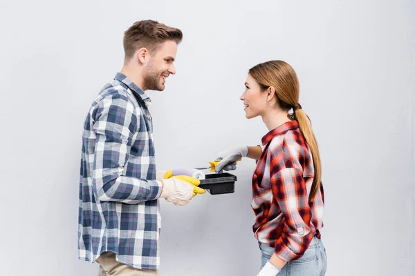 Vista lateral de feliz jovem casal com rolo de pintura e bandeja olhando um para o outro isolado no cinza — Fotografia de Stock