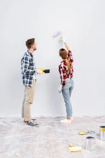 Pleine longueur de jeune femme avec mur de peinture à rouleaux près de l'homme tenant plateau à la maison — Photo de stock