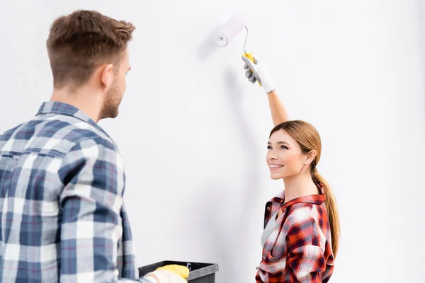 Feliz joven con la pared de la pintura del rodillo mientras que mira al hombre dentro - foto de stock