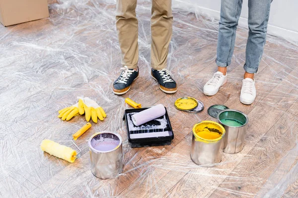 Cropped view of couple standing near tins of paint, gloves, tray and rollers on floor covered with polyethylene — Stock Photo
