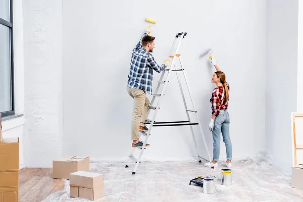 Longitud completa de sonriente pareja joven con rodillos pintando la pared y mirándose unos a otros cerca de la escalera en casa - foto de stock