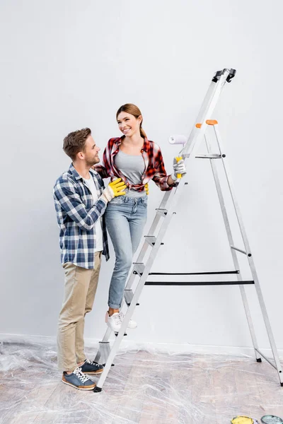 Longitud completa de sonriente hombre joven abrazando a la mujer con rodillo de pintura de pie en la escalera en casa - foto de stock