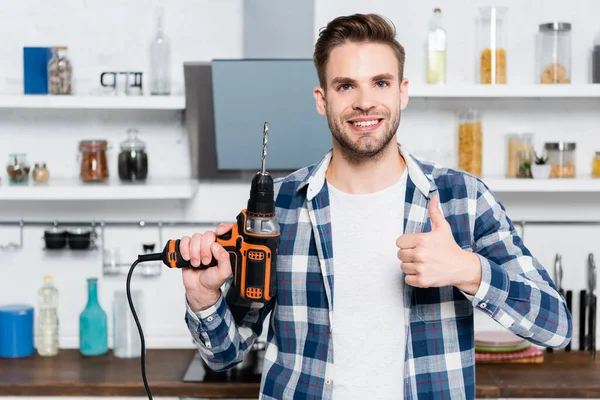 Front view of happy young man with drill looking at camera while showing thumb up with blurred kitchen on background — Stock Photo