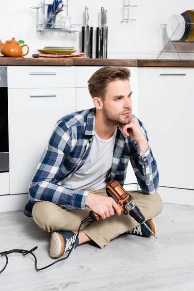 Longitud completa de joven hombre reflexivo con taladro mirando hacia otro lado mientras está sentado en el suelo en la cocina - foto de stock