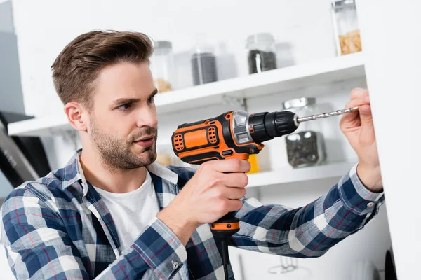 Focalizzato giovane uomo utilizzando trapano su sfondo sfocato in cucina — Foto stock