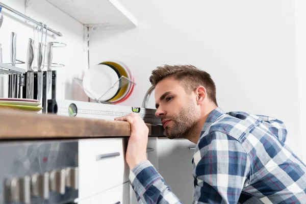 Концентрований молодий чоловік дивиться на інструмент вимірювання рівня на розмитому передньому плані на кухні — стокове фото