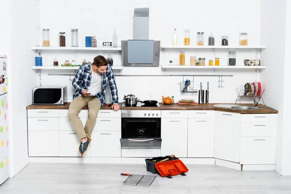 Comprimento total do jovem com tablet olhando para forno desmontado enquanto sentado na mesa na cozinha — Fotografia de Stock