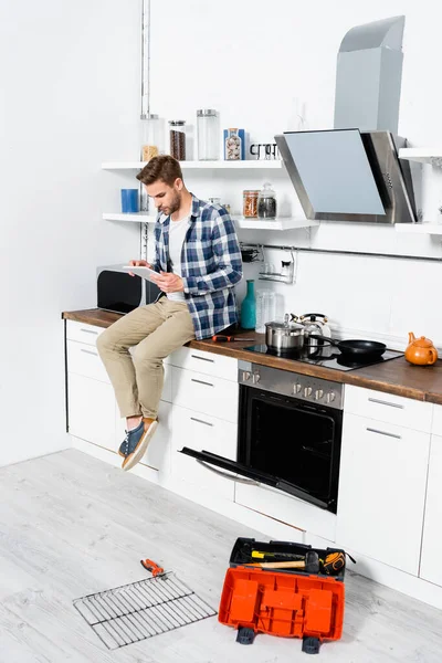 Intera lunghezza del giovane utilizzando tablet mentre seduto sul tavolo vicino al forno aperto e cassetta degli attrezzi in cucina — Foto stock