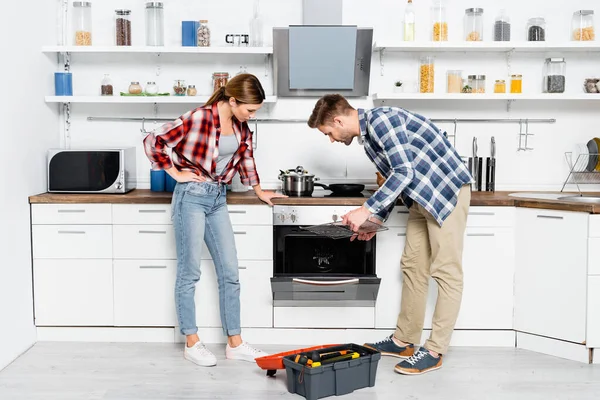 Tutta la lunghezza della giovane coppia piegandosi sopra il forno smontato e il toolkit aperto in cucina — Foto stock