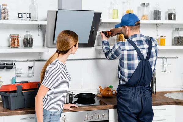 Visão traseira do faz-tudo com broca de reparação prateleiras perto de mulher na cozinha — Fotografia de Stock
