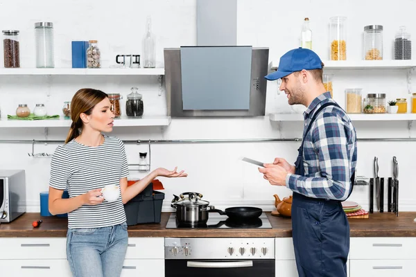 Mujer joven con taza de café hablando con manitas con tableta en la cocina - foto de stock