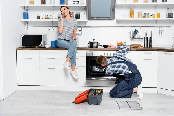 Volle Länge der lächelnden Frau, die auf dem Smartphone spricht, während sie auf dem Tisch neben dem Handwerker sitzt, der den Ofen in der Küche repariert — Stockfoto