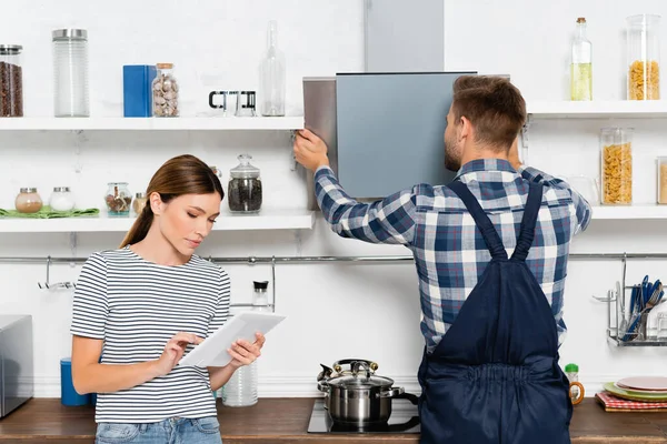 Young woman using tablet near handyman repairing extractor fan in kitchen — Stock Photo