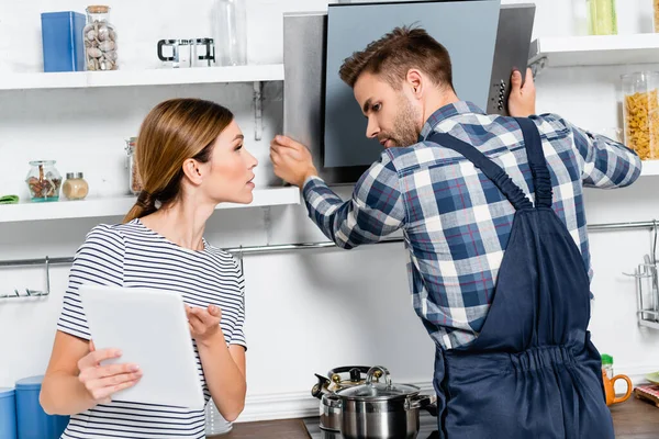 Young handyman looking at tablet while repairing extractor fan near woman in kitchen — Stock Photo