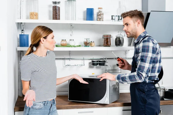 Young repairman with screwdriver looking at woman showing shrug gesture near microwave in kitchen — Stock Photo