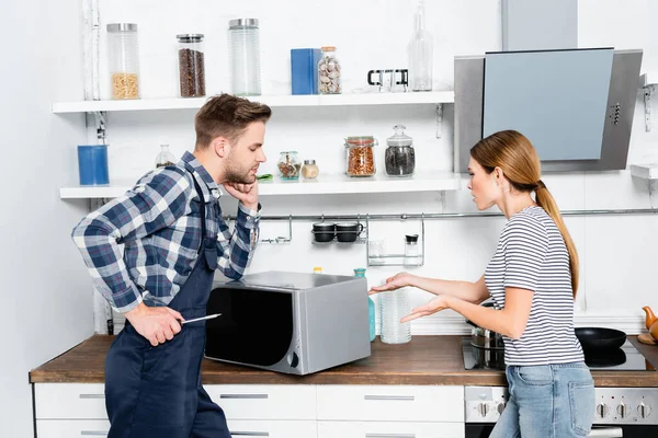 Junge Frau zeigt mit den Händen auf Mikrowelle in der Nähe von Reparateur mit Schraubenzieher in Küche — Stockfoto