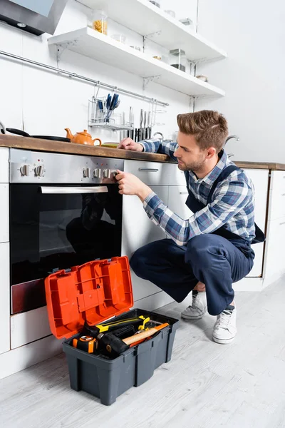 Junge Handwerker checken Backofenknopf, während sie in der Küche neben Werkzeugkiste sitzen — Stockfoto