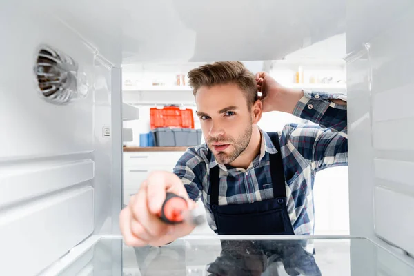 Jeune réparateur réfléchi avec tournevis regardant la caméra dans le congélateur sur le premier plan flou — Photo de stock