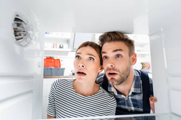 Shocked young woman and handyman with open mouths looking at freezer on blurred foreground in kitchen — Stock Photo