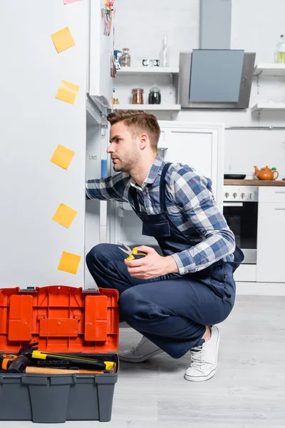 Longitud completa de reparador joven con alicates fijación nevera mientras está sentado en el piso cerca de la caja de herramientas abierta en la cocina - foto de stock