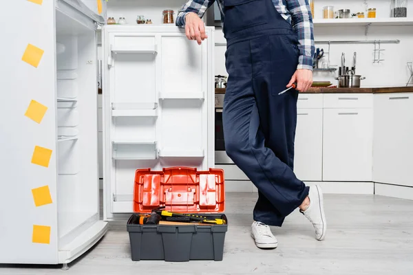 Ausgeschnittener Blick auf jungen Handwerker mit Schraubenzieher an Kühlschrank in der Nähe von Werkzeugkiste in Küche gelehnt — Stockfoto