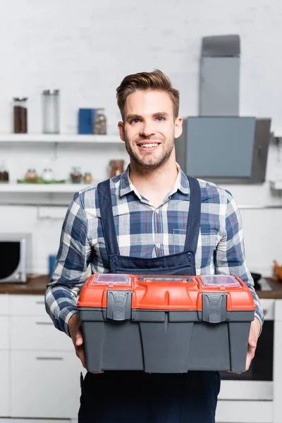 Vista frontal do reparador feliz olhando para a câmera enquanto segurando caixa de ferramentas com cozinha embaçada no fundo — Fotografia de Stock