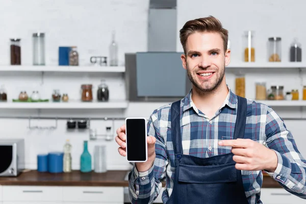 Visão frontal do jovem feliz olhando para a câmera ao apontar com o dedo para o smartphone no fundo borrado na cozinha — Fotografia de Stock