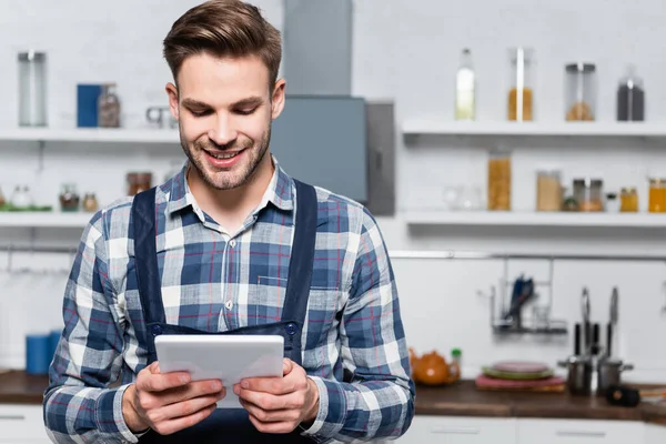 Vue de face de heureux bricoleur tenant tablette avec cuisine floue sur fond — Photo de stock