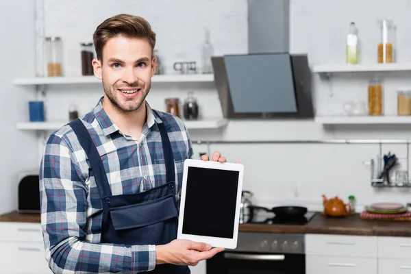 Front view of happy handyman looking at camera while showing tablet with blurred kitchen on background — Stock Photo