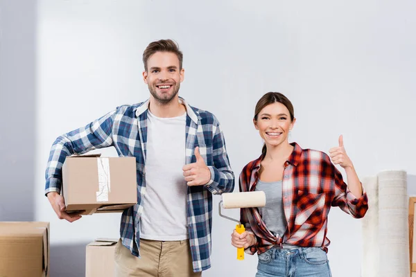 Frontansicht eines glücklichen jungen Paares mit Schachtel und Farbroller, die Daumen nach oben zeigen, während sie drinnen in die Kamera schauen — Stockfoto