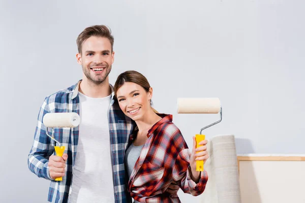 Vue de face du jeune couple heureux avec des rouleaux de peinture regardant la caméra tout en étreignant à la maison — Photo de stock