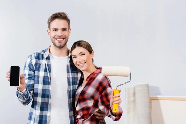 Vista frontal de la joven pareja sonriente con rodillo de pintura y teléfono inteligente mirando a la cámara mientras se abraza en casa - foto de stock
