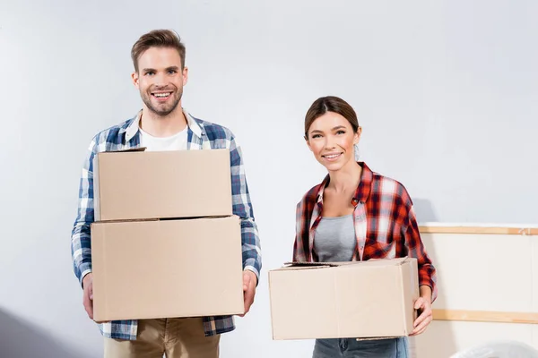 Frontansicht eines fröhlichen jungen Paares, das in die Kamera schaut, während es zu Hause Kartons hält — Stockfoto