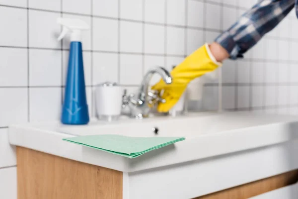 Rag sur évier près du détergent et femme près du robinet sur fond flou dans la salle de bain — Photo de stock