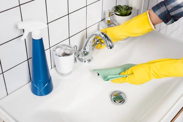 Vista ritagliata della donna in guanti di gomma straccio bagnante vicino detergente sul lavandino in bagno — Foto stock